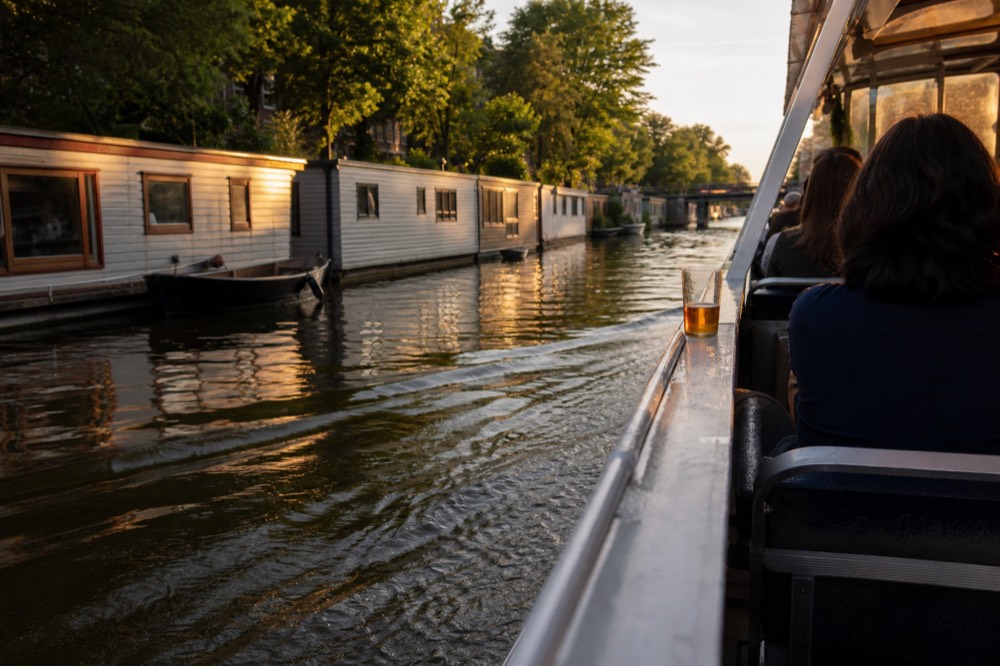 A canal cruise boat passing a row of houseboats on an Amsterdam canal at golden hour, no text, no logos, no people