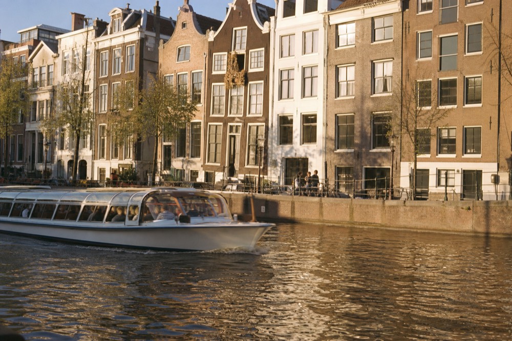 Canal cruise boat passing gabled houses on a wide Amsterdam canal in soft evening light, no text, no logos, no people