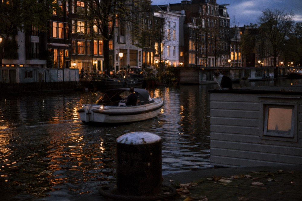 A small saloon boat cruising past illuminated Amsterdam canal houses at dusk, no text, no logos, no people