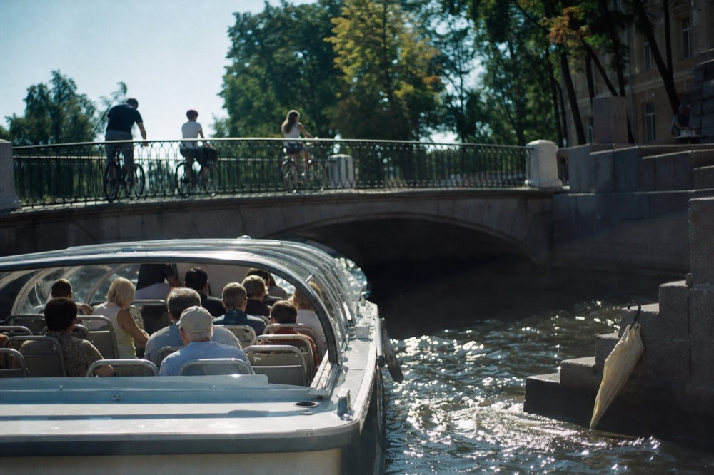 An Amsterdam canal cruise boat approaching a low arched bridge with cyclists on the quay above, no text, no logos, no people