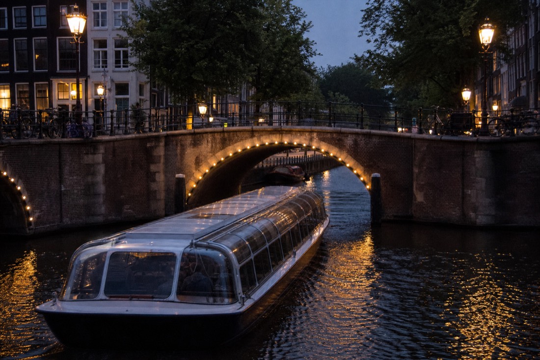 A floodlit Amsterdam canal bridge at dusk with a dinner cruise boat passing beneath, warm reflections on dark water, no text, no logos, no people