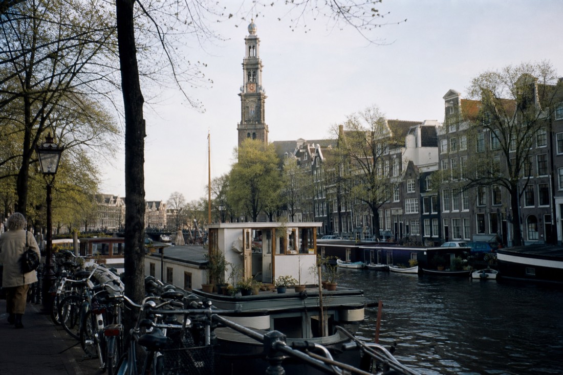 Row of houseboats moored along the Prinsengracht with the Westerkerk tower behind, no text, no logos, no people.