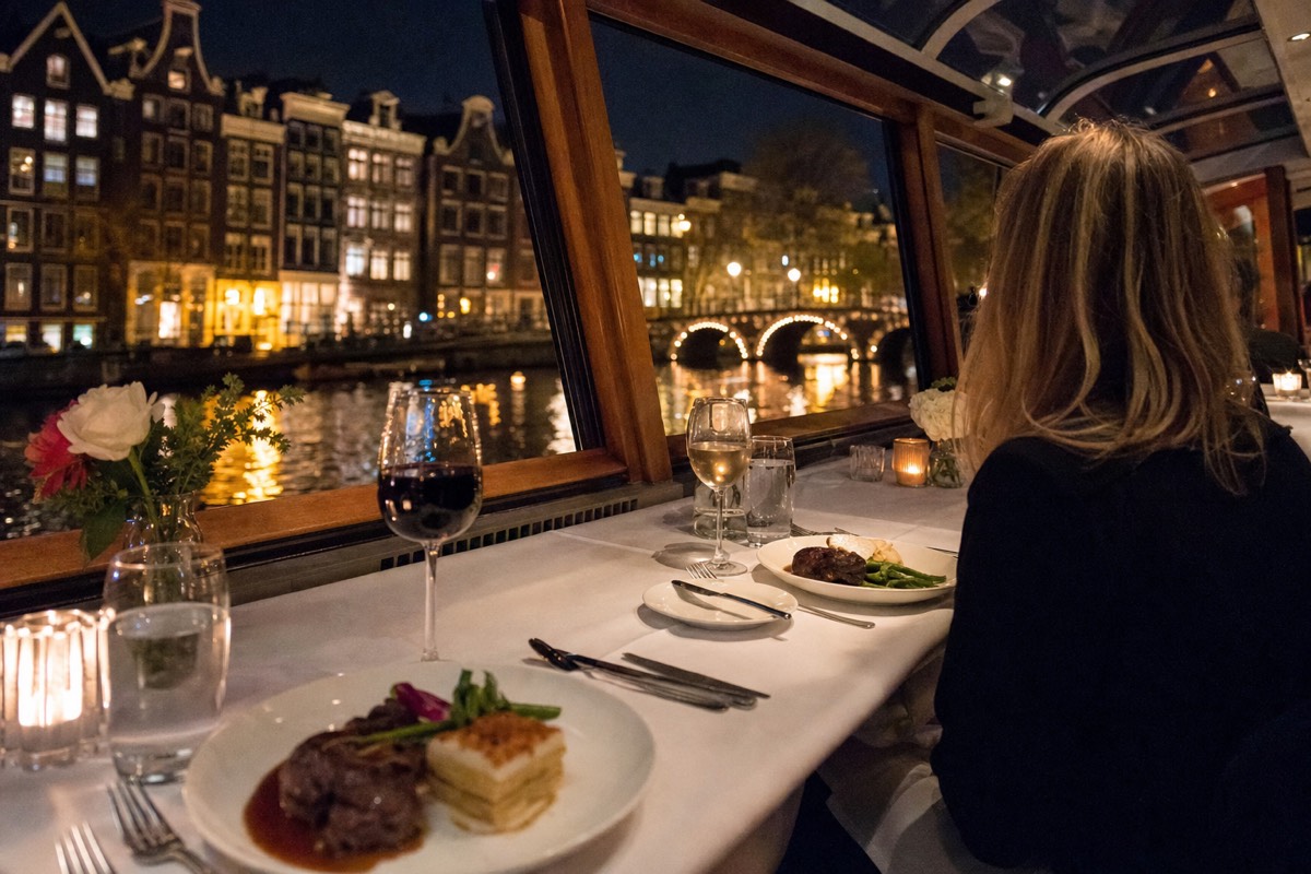 A candlelit dinner table set with wine glasses and plated dishes beside a canal boat window overlooking illuminated Amsterdam canal houses and a lit bridge at night