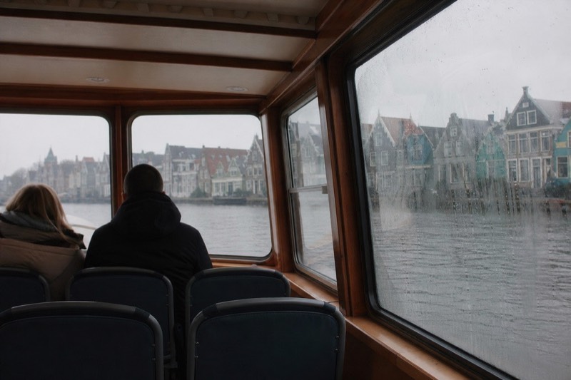 Passengers seen from behind aboard a small, uncrowded heated saloon boat gliding past gabled Amsterdam canal houses, wood-paneled interior and empty seats visible through the windows on a quiet afternoon