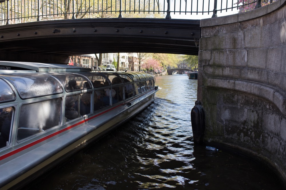 A glass-topped canal cruise boat passing under a low Amsterdam bridge with sightseers absent from the frame, no text, no logos, no people