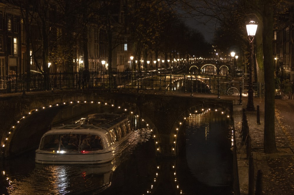 An Amsterdam canal cruise boat at night with warm lit bridges reflecting on black water, no text, no logos, no people