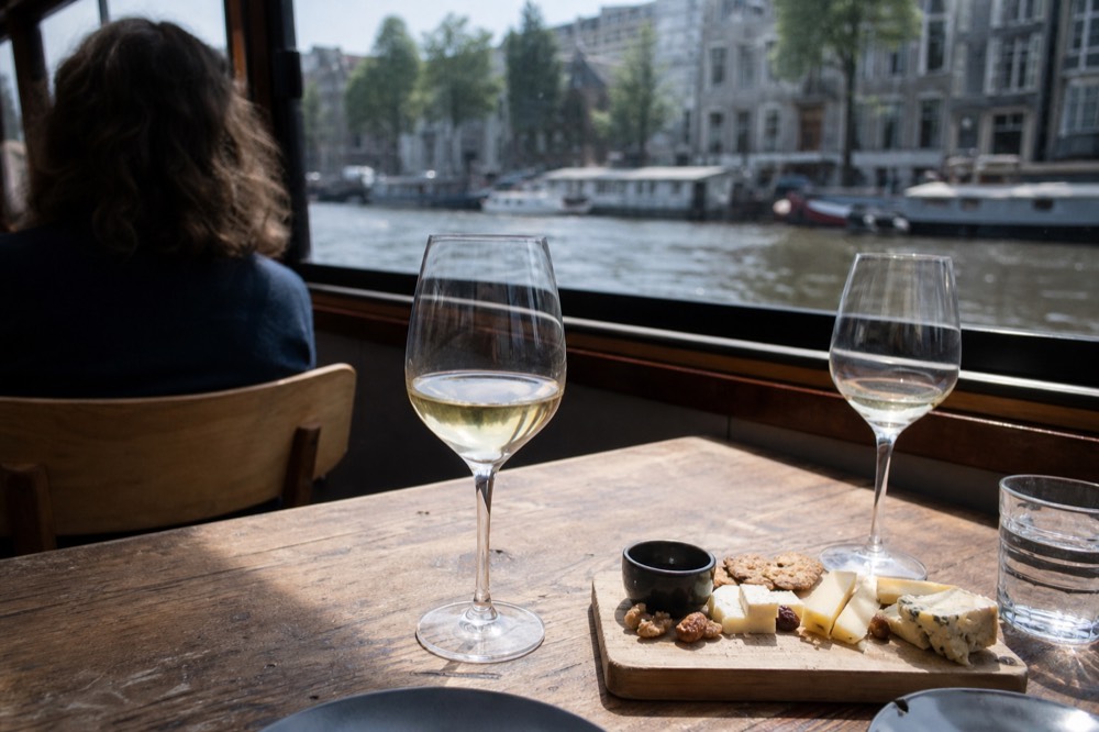 Glasses of white wine and a cheese board set on a wooden canal boat table with canal houses blurred behind, no text, no logos, no people