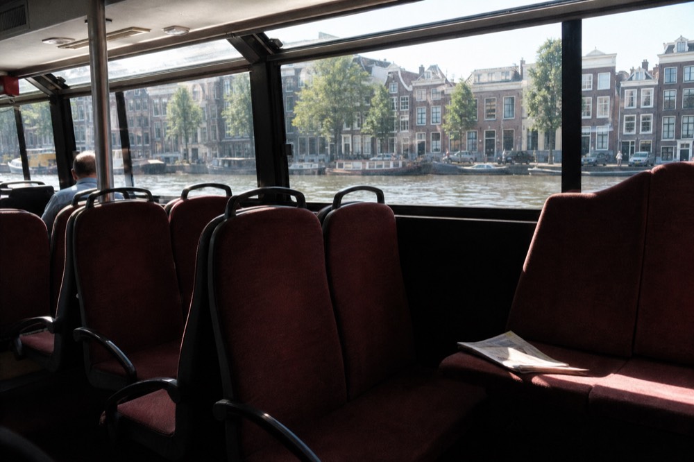 Rows of seats inside a covered canal cruise boat with large windows facing Amsterdam canal houses, no text, no logos, no people