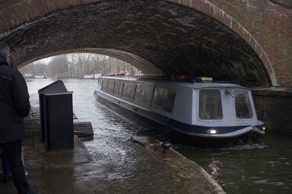 A canal boat passing under a low arched Amsterdam bridge with the fare and boarding area visible at the quay, no text, no logos, no people