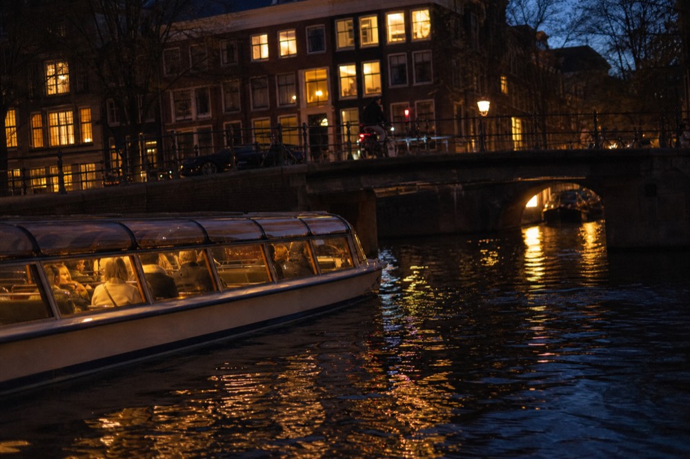 An evening canal cruise boat lit from inside passing illuminated Amsterdam canal houses after dark, no text, no logos, no people