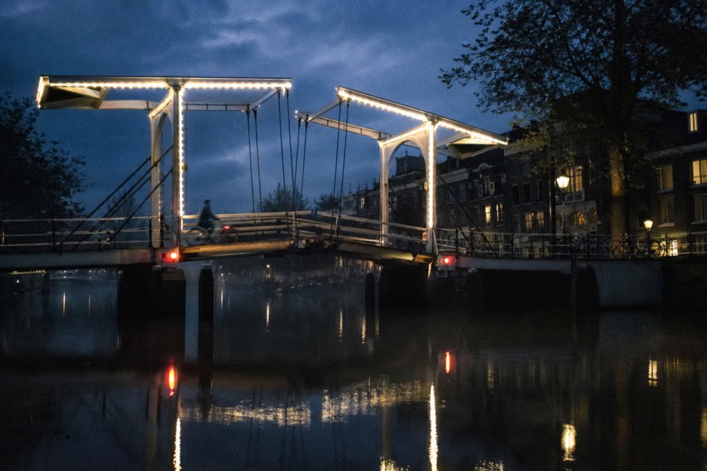 Illuminated Amsterdam bridge reflecting in dark canal water at dusk, no text, no logos, no people