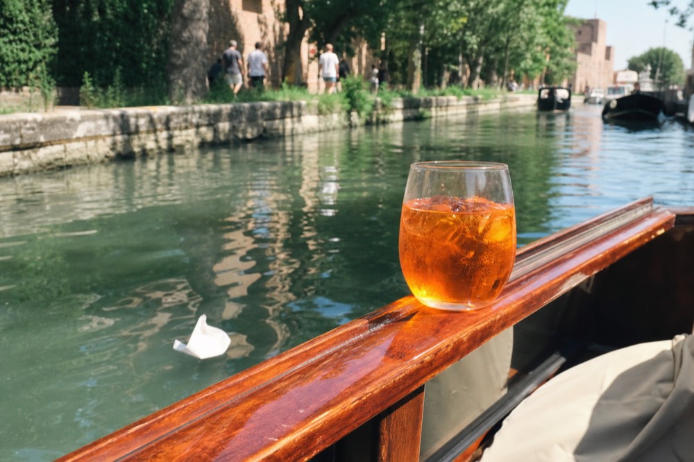 An orange spritz cocktail on a boat rail with canal reflections, no text, no logos, no people.