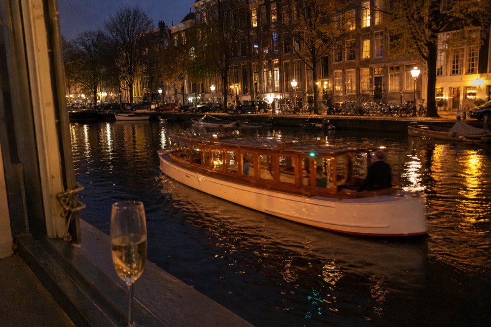 An elegant salon boat cruising past illuminated Amsterdam canal houses in the evening, no text, no logos, no people