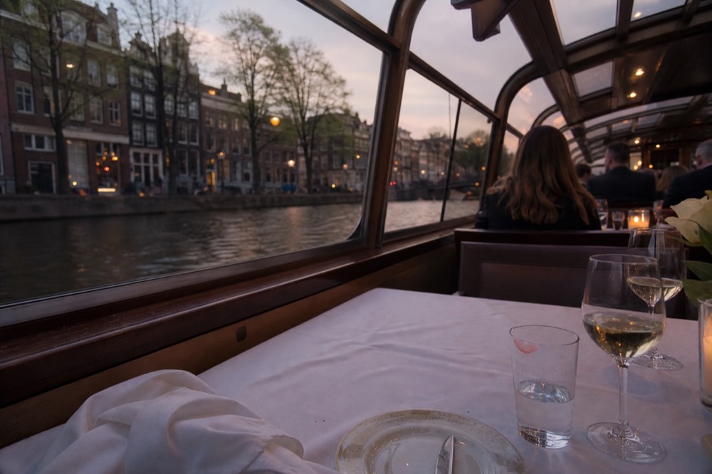 A set dinner table with white linen inside a covered canal boat, canal houses visible through the window, no text, no logos, no people.