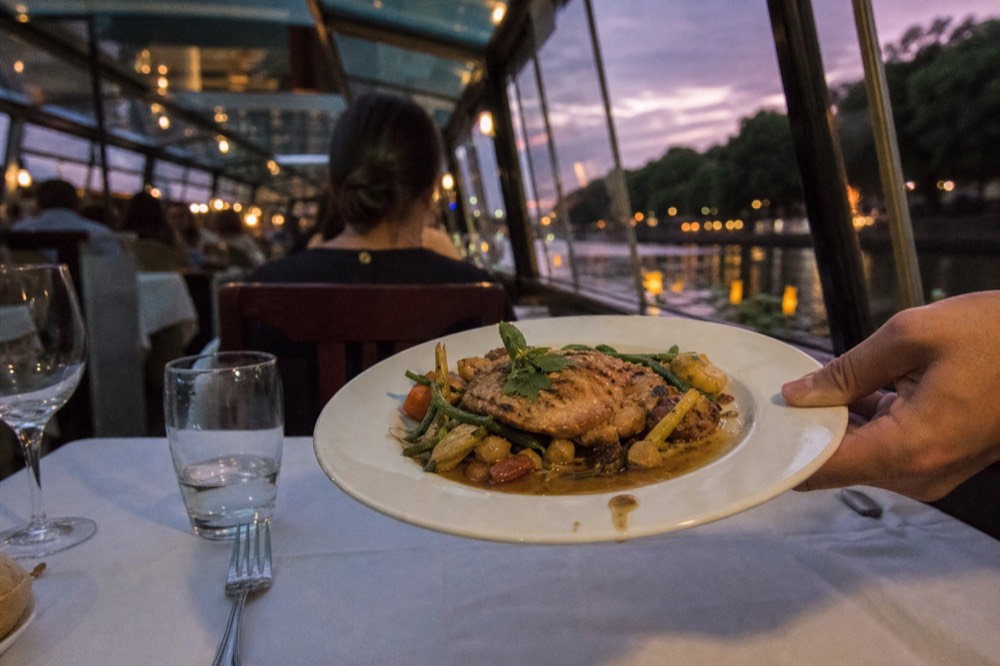 A plated main course being served at a table inside a glass-covered canal cruise boat at dusk, no text, no logos, no people.