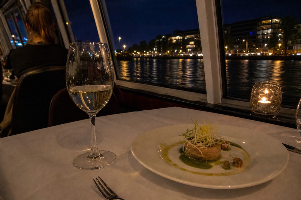 A glass of white wine and a plated starter on a dinner table aboard a canal boat, reflections of canal lights in the window, no text, no logos, no people.