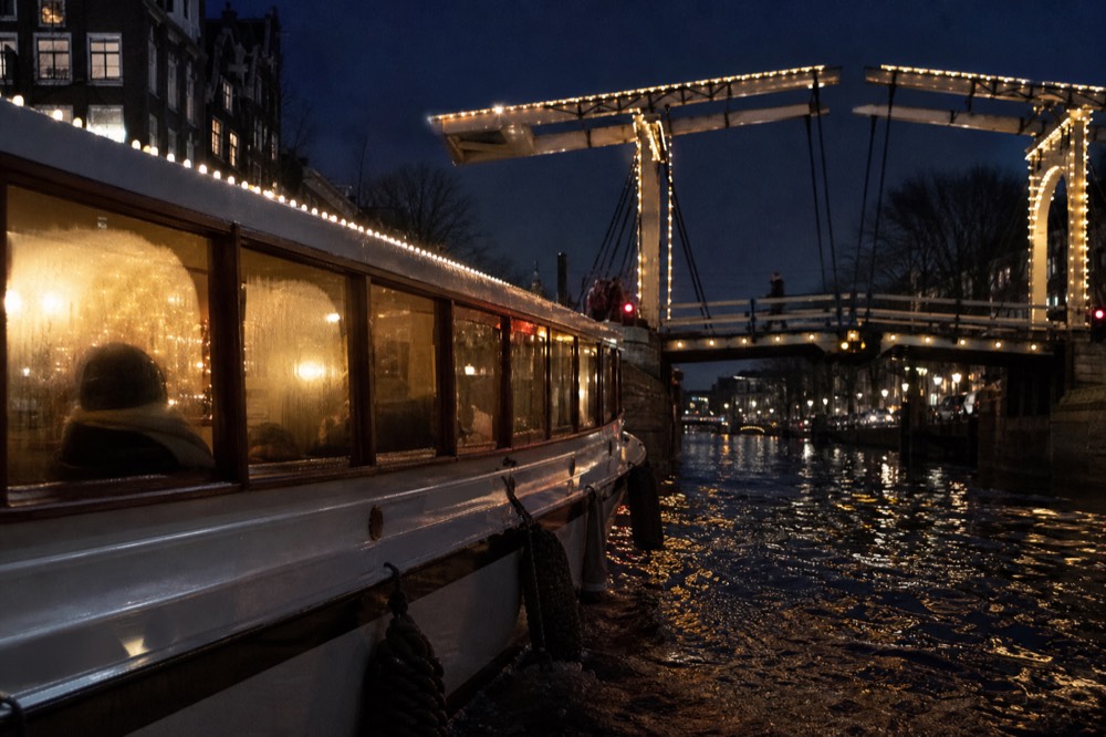 A canal cruise boat passing under a lit bridge in Amsterdam at night, seen from the water, no text, no logos, no people.