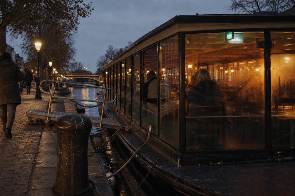 A covered dinner cruise boat moored at a jetty on an Amsterdam canal at dusk, warm light inside the cabin, no text, no logos, no people.