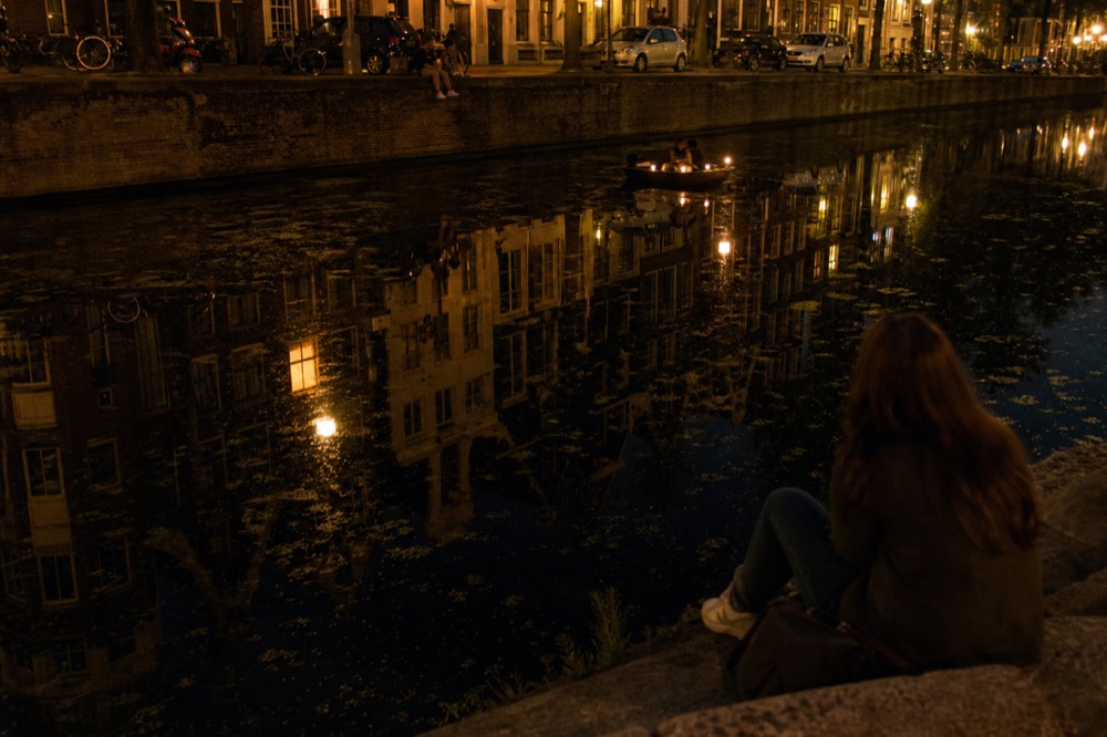 Amsterdam canal houses reflected in dark water at night with a small cruise boat's lights in the distance, no text, no logos, no people.