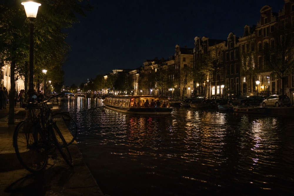 A wide view of an Amsterdam canal at night with a lit cruise boat passing gabled houses, no text, no logos, no people.