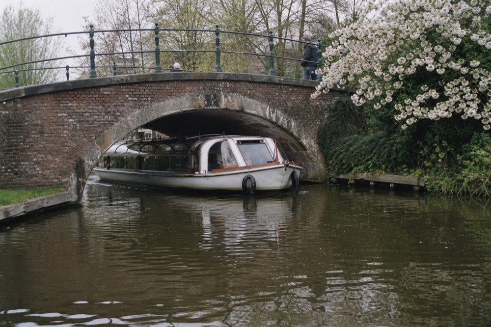 A classic glass-topped canal cruise boat passing under a low Amsterdam bridge, no text, no logos, no people