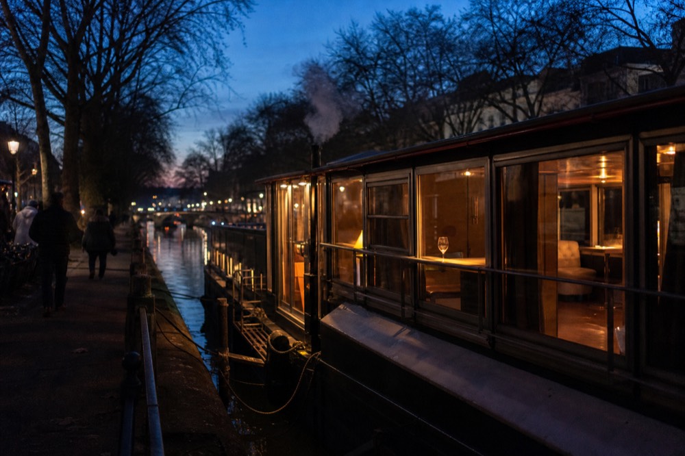 A dinner cruise boat lit from inside, moored beside an evening canal in Amsterdam, no text, no logos, no people