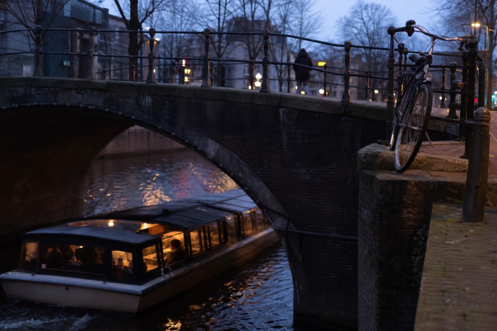 A covered canal boat gliding under a low Amsterdam bridge at dusk, no text, no logos, no people